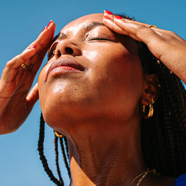 Woman with sun-kissed skin holding her forehead under bright sun, showing signs of heat or sunburn. Wearing gold jewelry and red nail polish, against a clear blue sky
